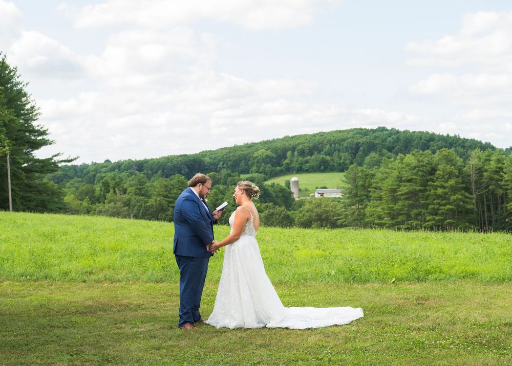 A couple stands together in an open green meadow surrounded by rolling hills and trees on their wedding day.