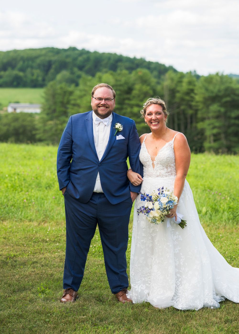 Newlywed couple poses playfully in a green summer meadow wearing navy suit and white wedding gown with bouquet.