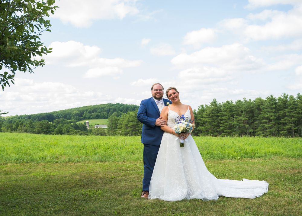 Wedding couple poses in scenic field with rolling hills and trees in background.