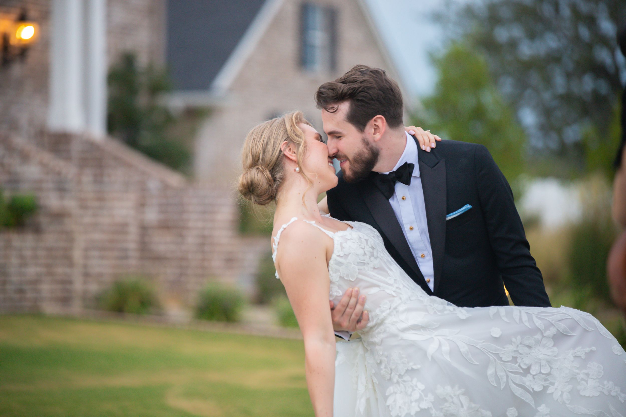 A romantic wedding couple shares a tender embrace on the lawn of a brick building.