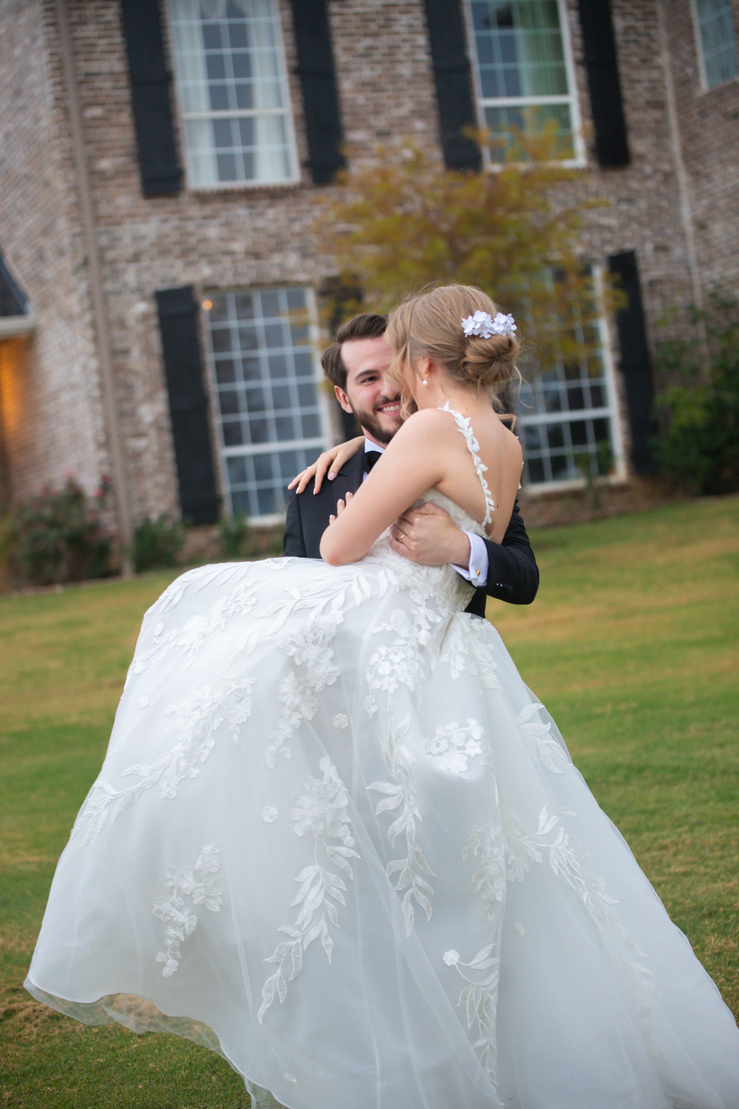 A couple in wedding attire dances and spins on a green lawn near a historic brick building.