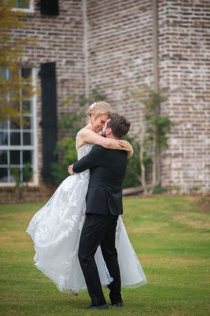Wedding couple shares a playful dance moment on the grass near a brick building.