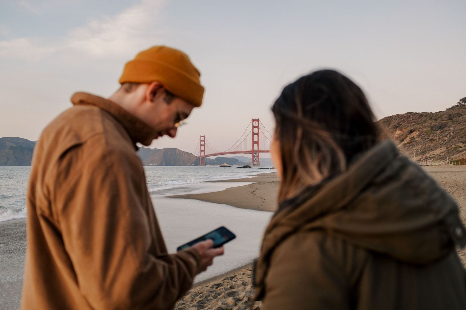 Two people looking at the Golden Gate Bridge from Baker Beach during sunset.