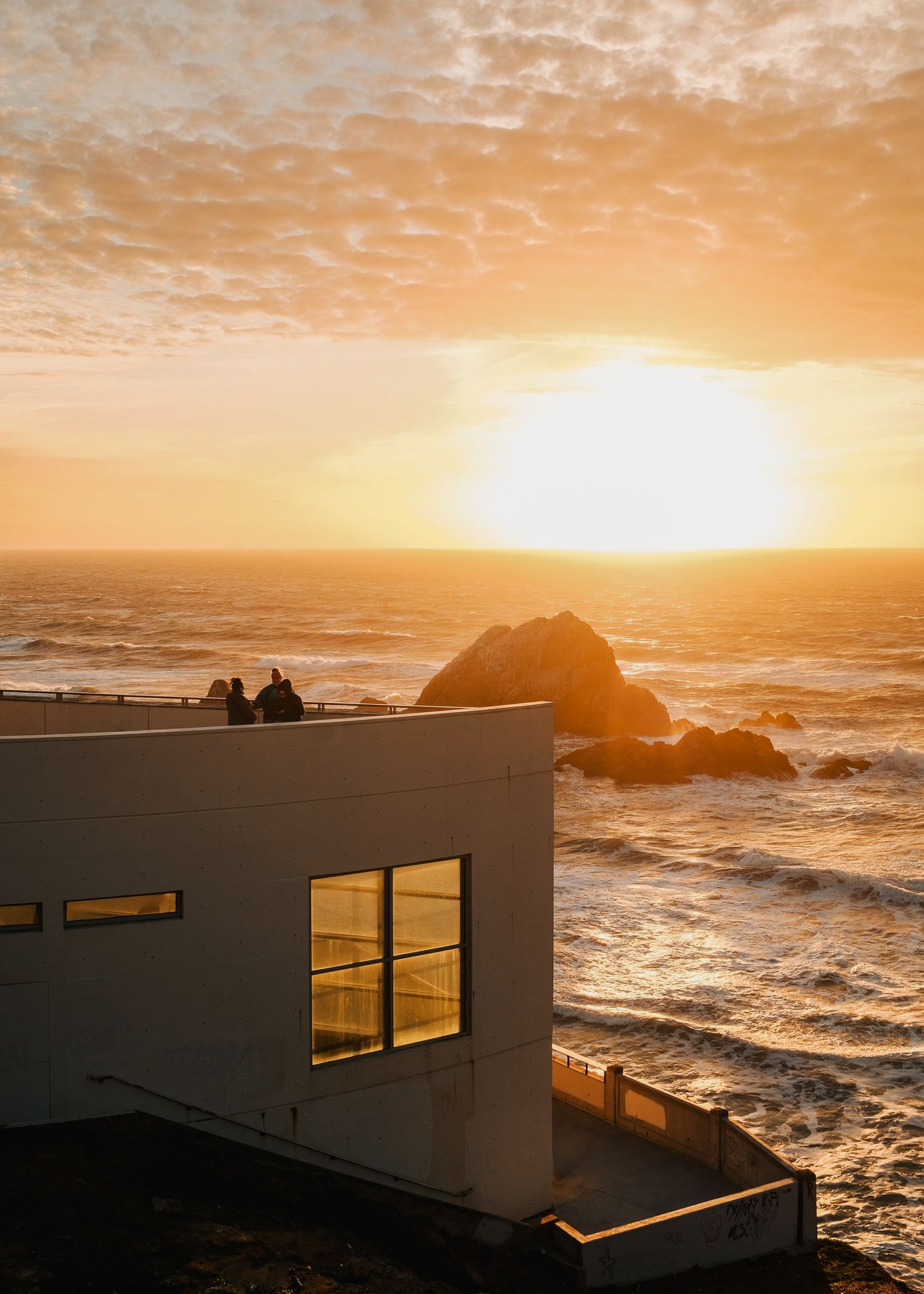 White coastal building overlooking dramatic ocean sunset with waves crashing against rocky shore.
