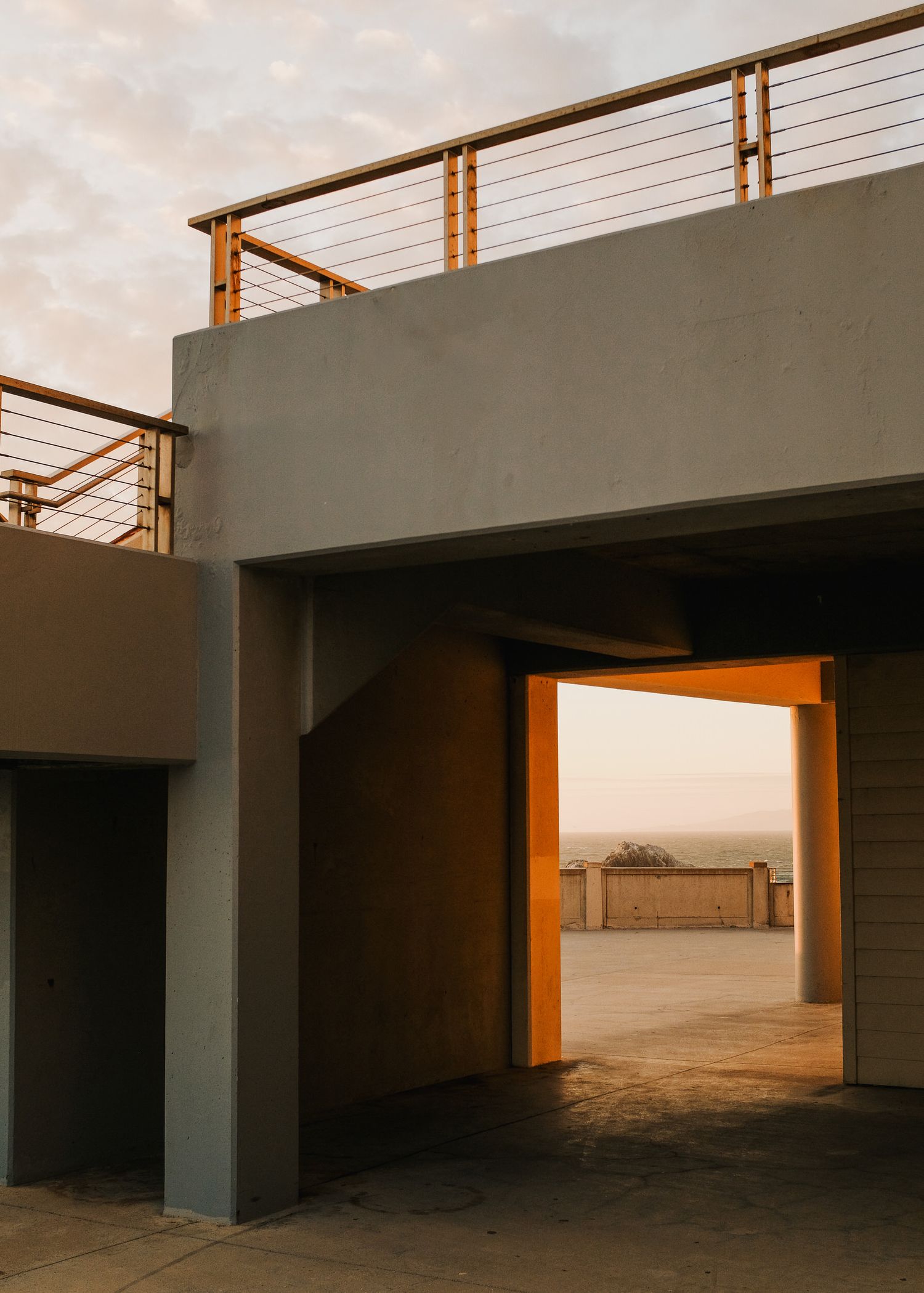 Modern architectural passageway with concrete walls and wooden railings illuminated by warm sunset light through an orange doorway.