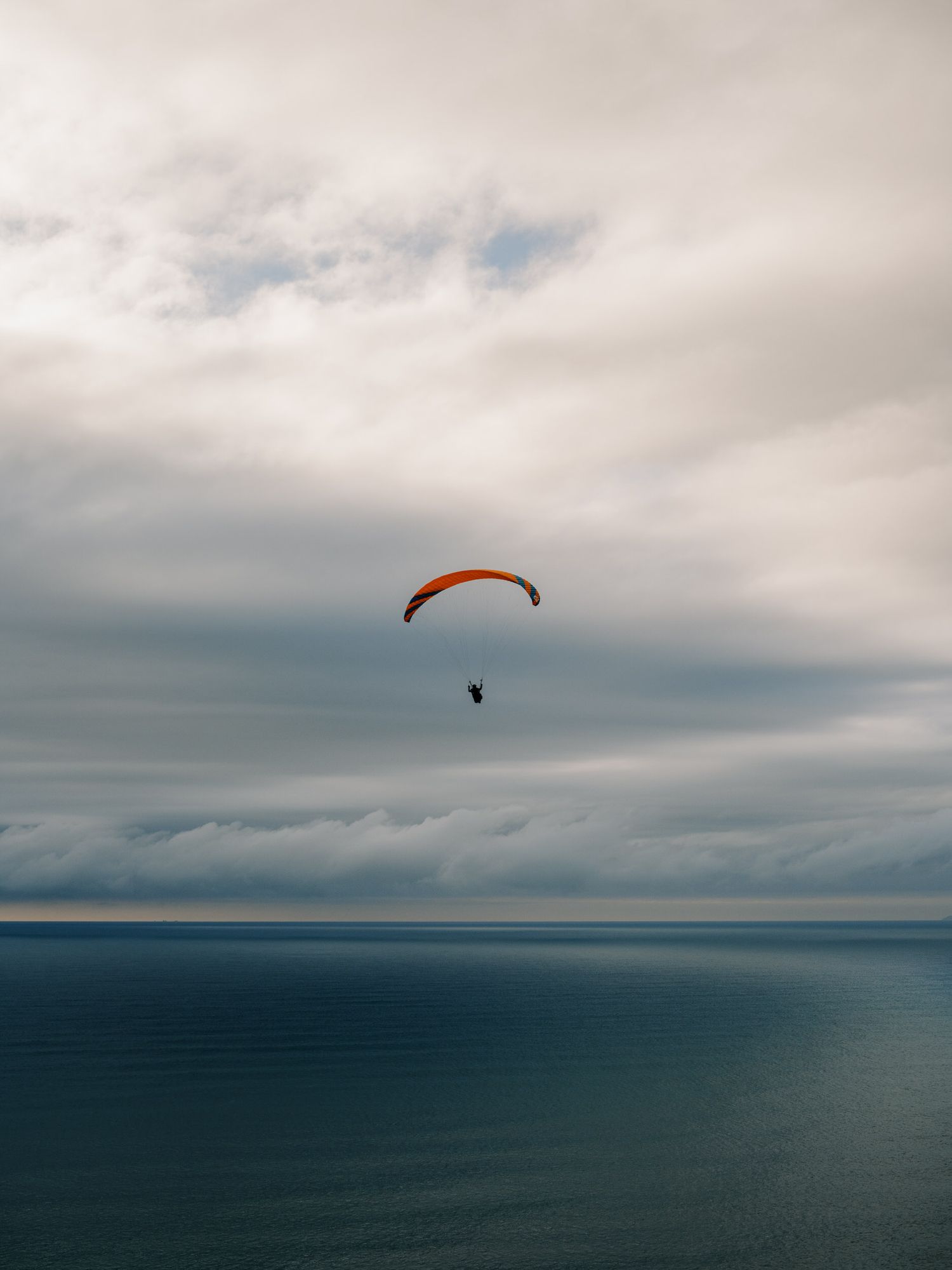 Orange paraglider floating against moody cloudy sky over dark ocean waters.