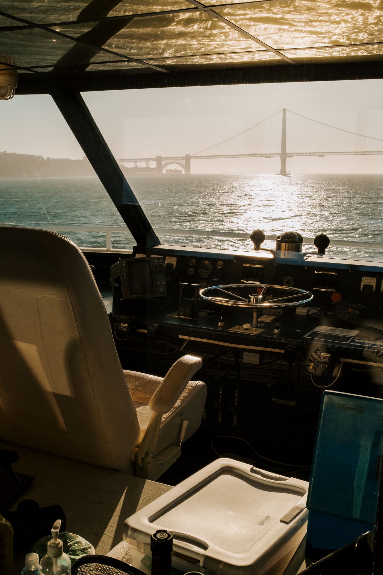 View of Golden Gate Bridge through boat cockpit window with captain's chair and control panel at sunset.
