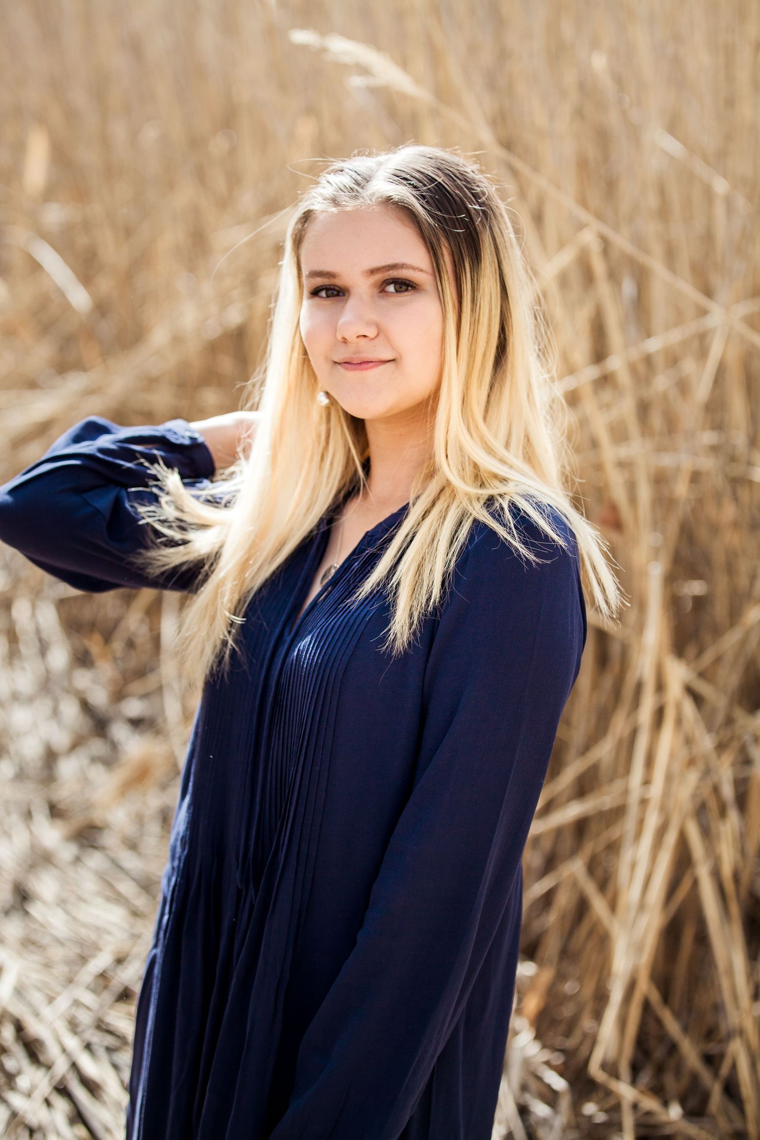 A person in a blue top poses outdoors against a backdrop of tall dried wheat or grass stalks on a sunny day. Graduation Portrait. Slc Utah.