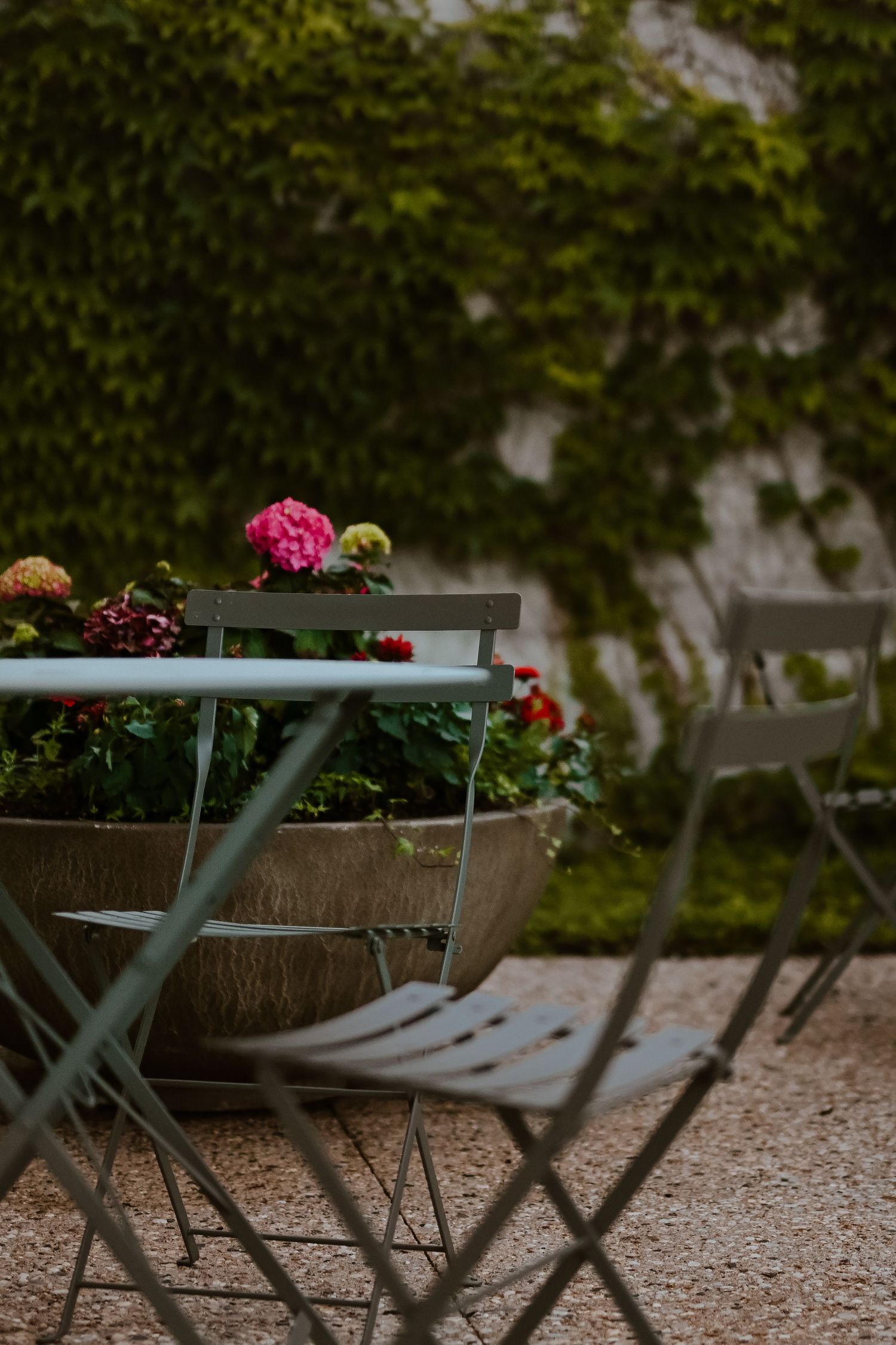 Sage green bistro chairs and table on gravel patio beside a large planter with pink flowers, ivy-covered wall backdrop.