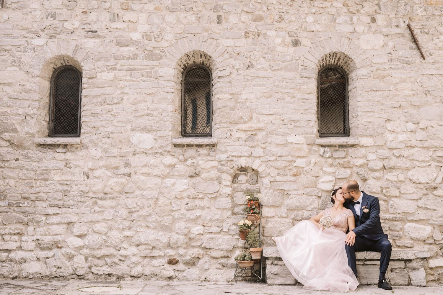 Coppia di sposi che si baciano seduti su una panchina di sasso all'esterno di una chiesa