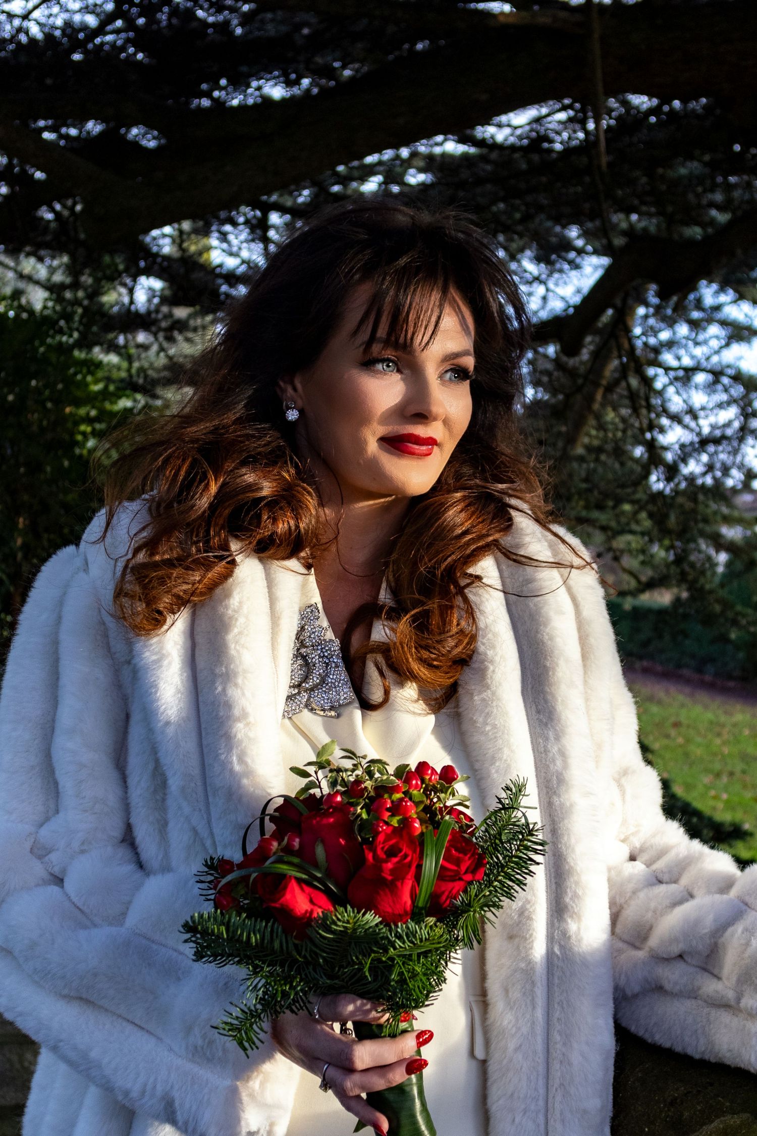 Winter bride in white fur coat holds red bouquet against evergreen backdrop.