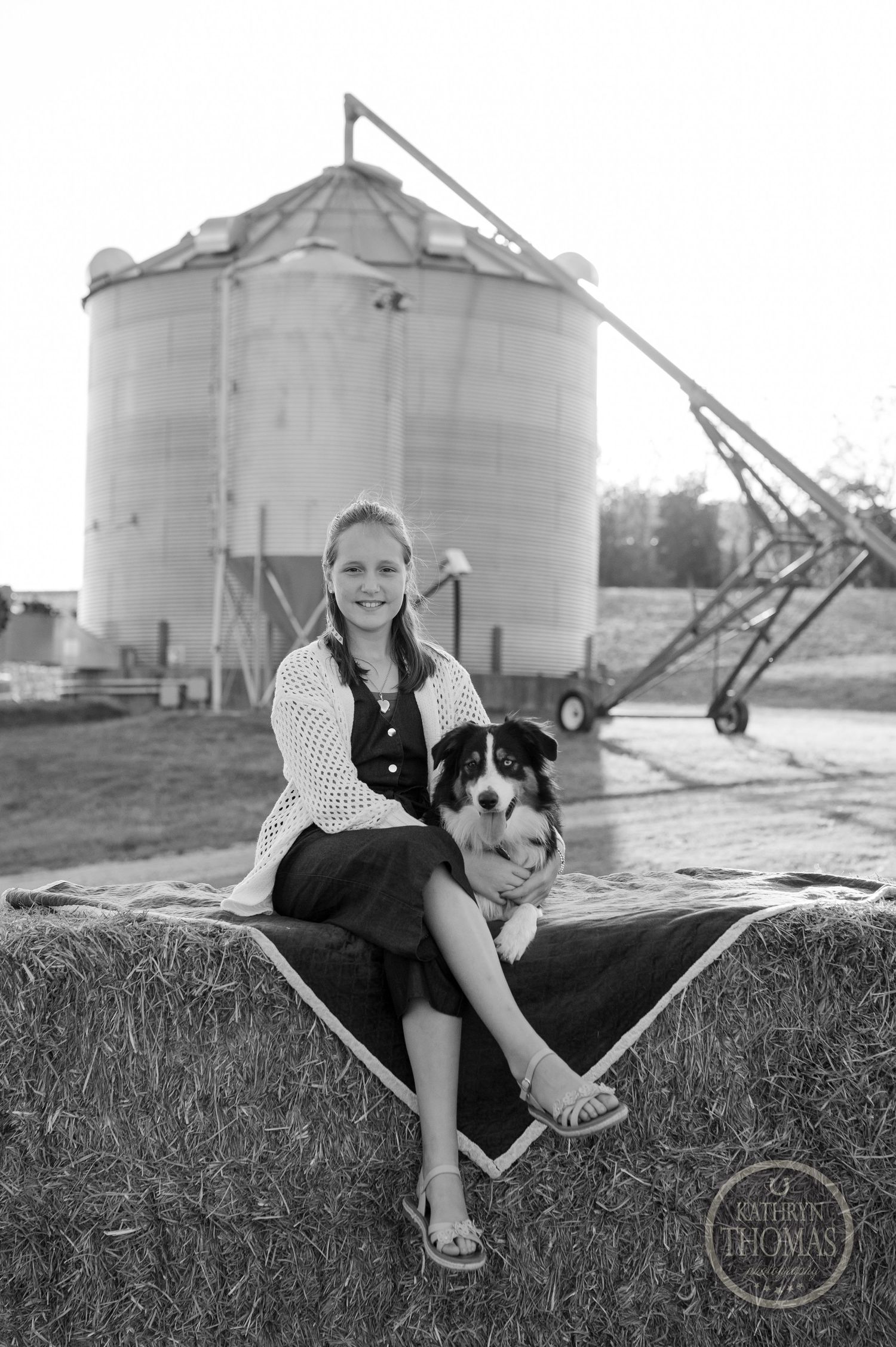 Black and white portrait of person with Australian Shepherd on hay bale near grain silo.