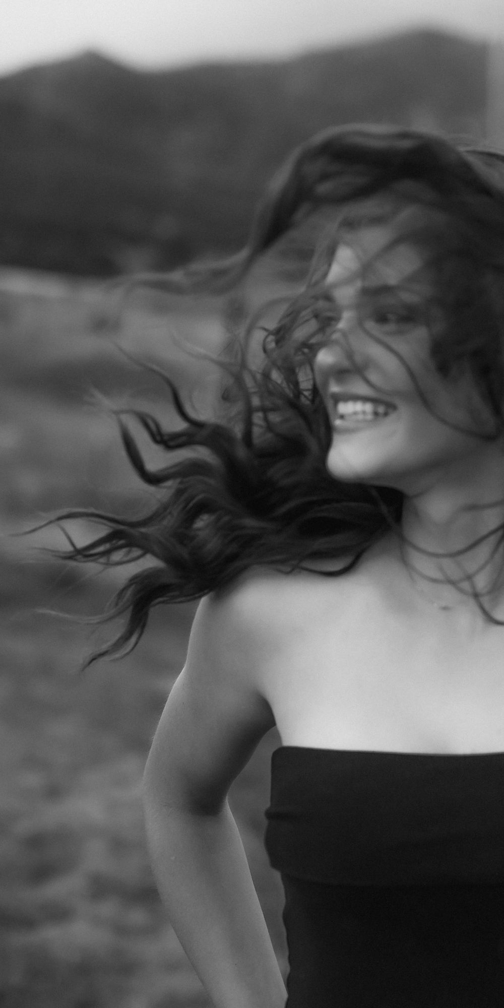 Black and white portrait of woman with windswept hair laughing against mountain backdrop.