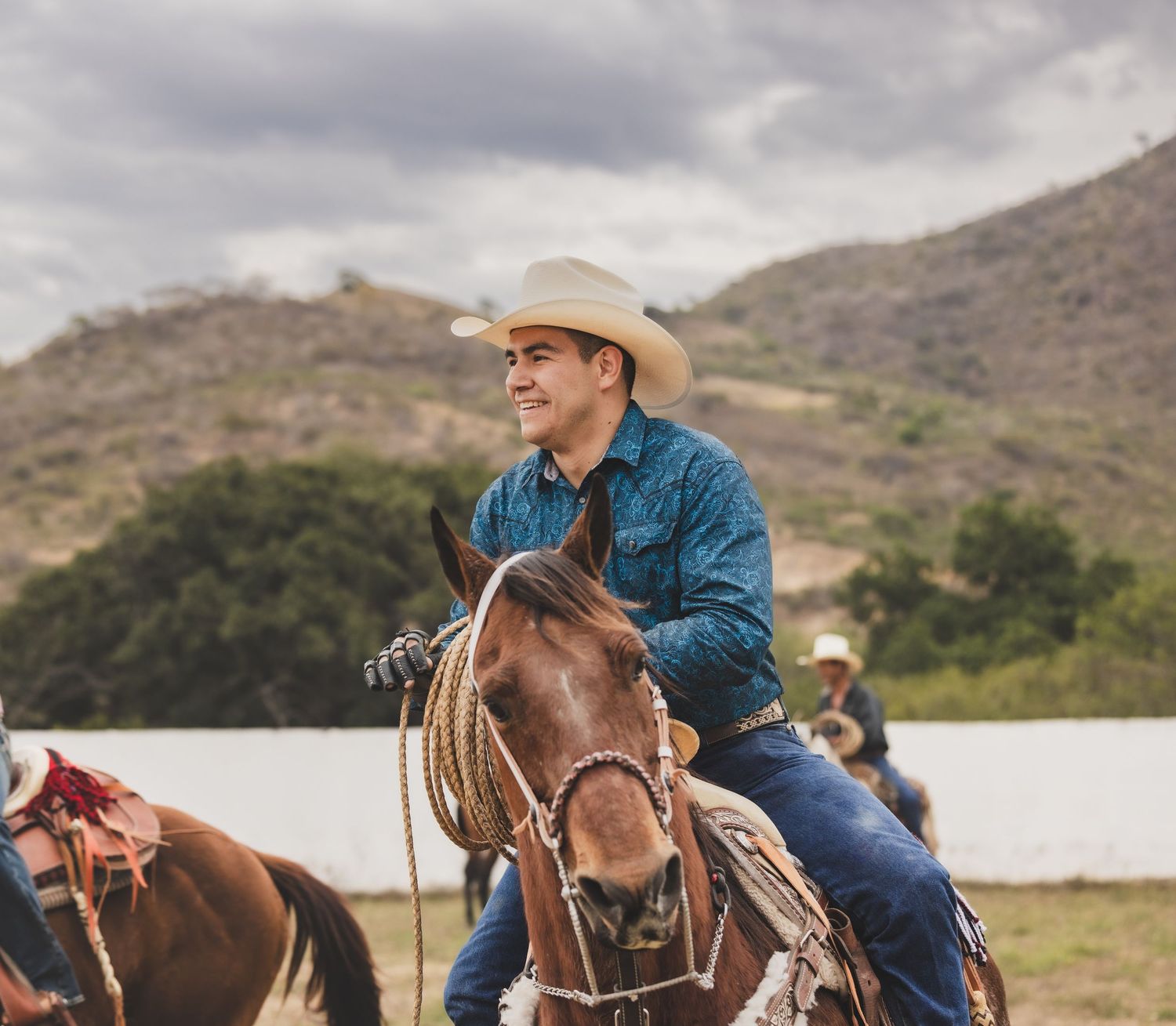 Smiling cowboy in straw hat riding a horse with lasso at a rodeo with hills behind.