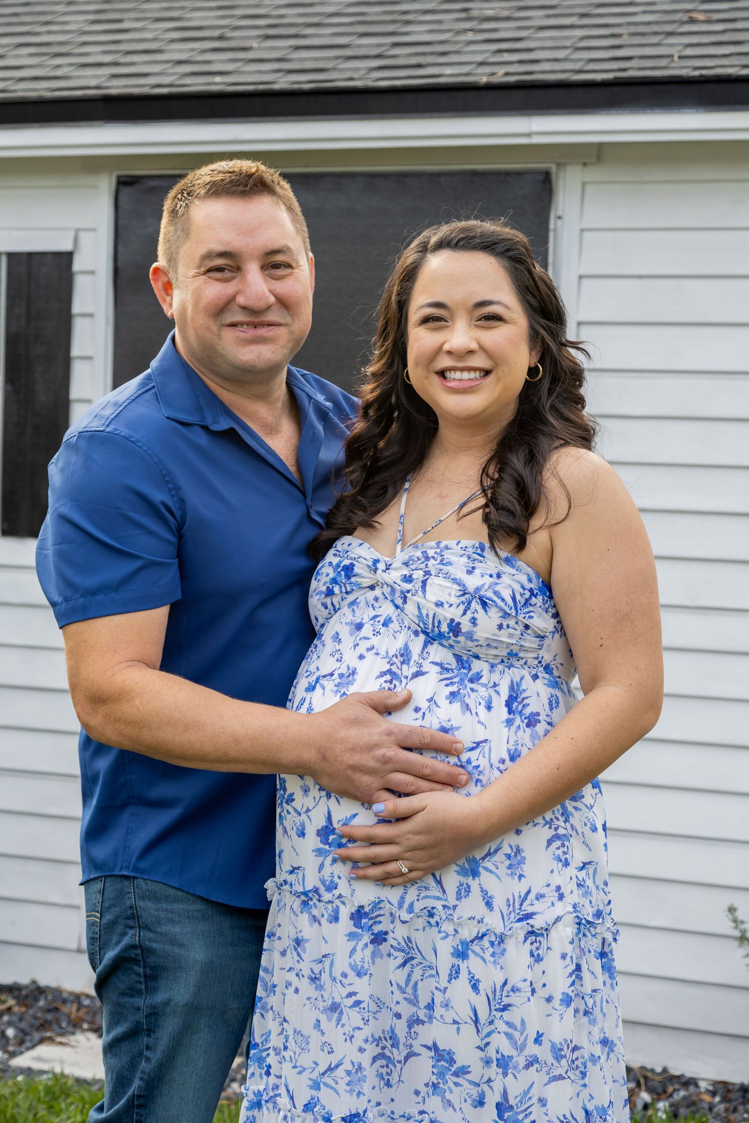 Smiling couple posing outdoors, man in blue polo embracing pregnant woman in blue floral maternity dress.