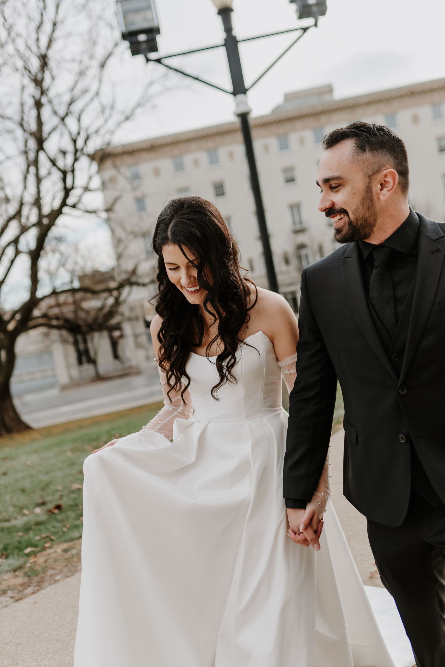 Newlywed couple walking outdoors with bride in strapless white dress and groom in black suit holding hands.