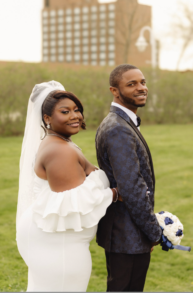 A couple in wedding attire poses back to back on a grassy lawn with city buildings in the background.