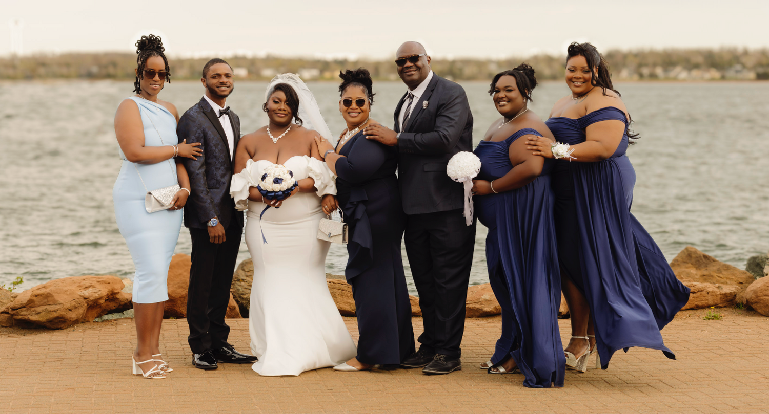 Wedding party in formal attire poses together on rocky shoreline with water and sky in background.