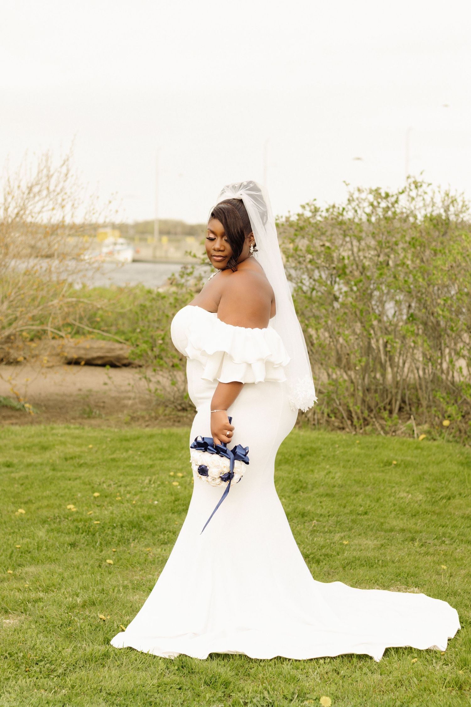A bride in an elegant off-shoulder mermaid wedding dress poses on a grassy lawn holding a navy and white bouquet.