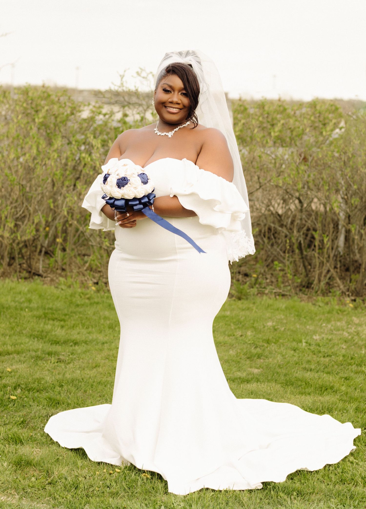 Beautiful bride in a fitted white mermaid wedding gown holds a blue and white bouquet while standing in a garden setting.