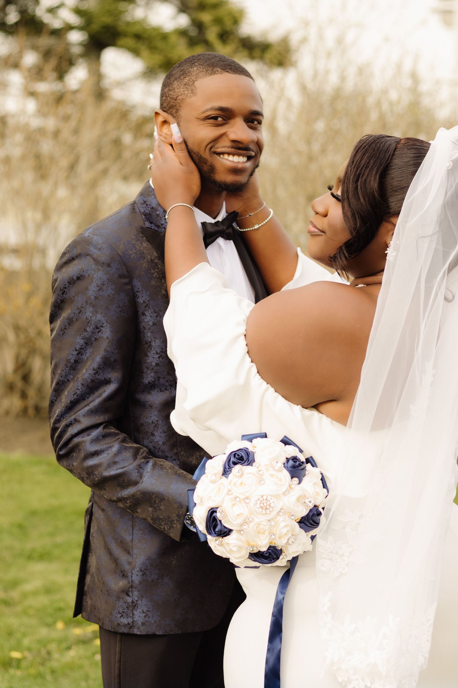 Newlywed couple in formal wedding attire share a tender moment outdoors while holding a navy and white floral bouquet.