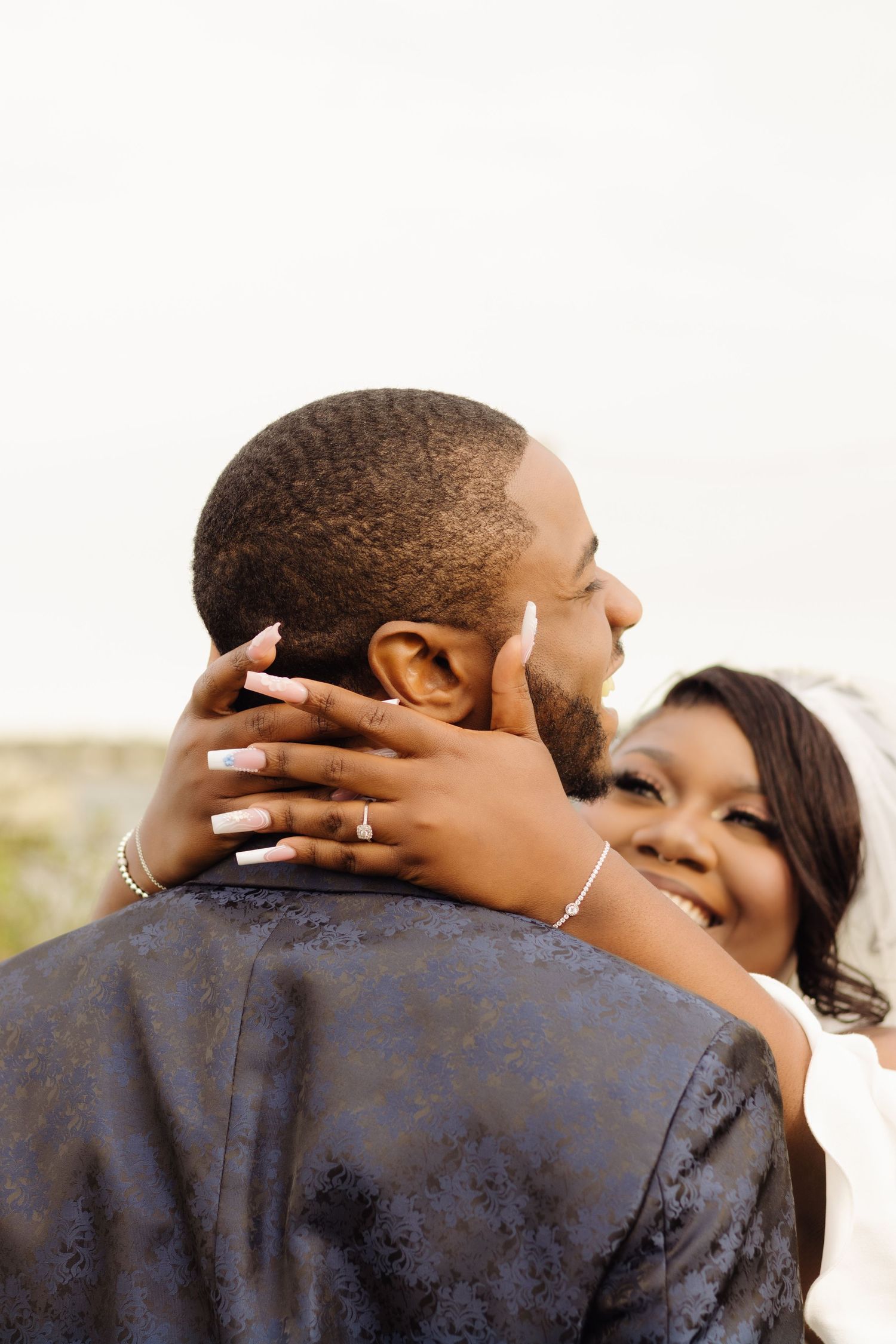 A couple shares an intimate embrace outdoors, with delicate gold rings and jewelry visible on their hands.