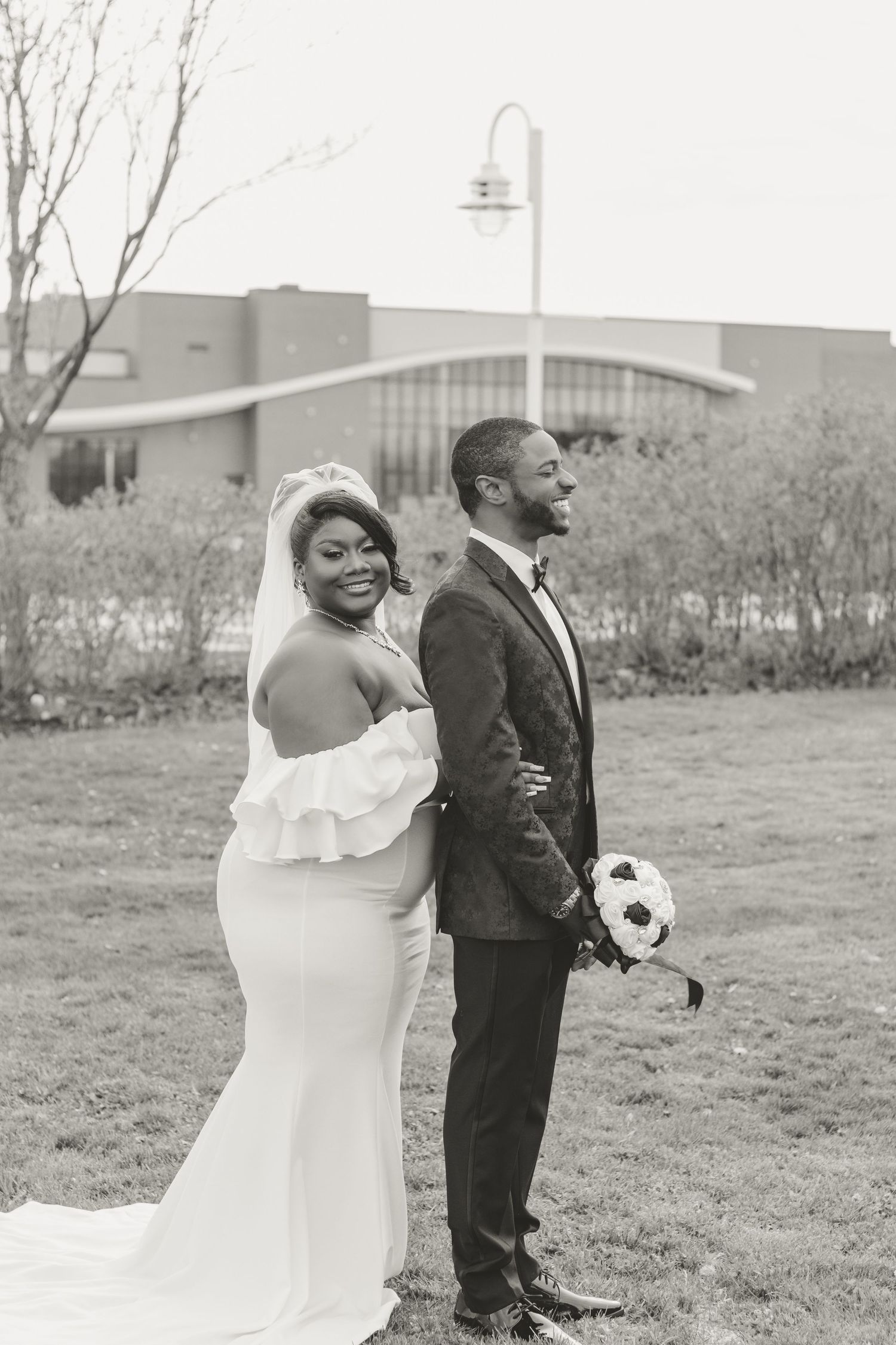 A happy couple stands together in a romantic black and white wedding photo outdoors with urban buildings in background.