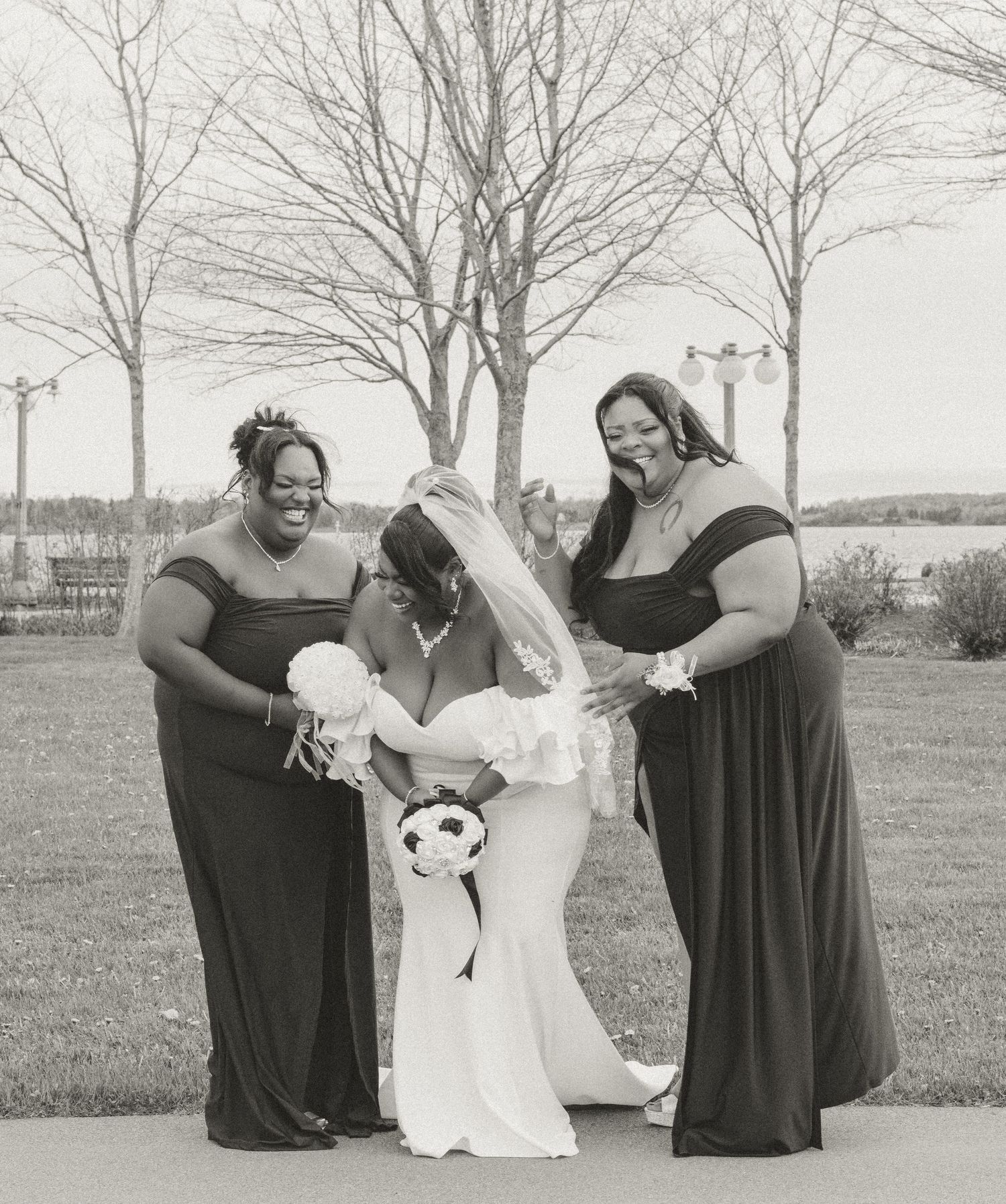 Black and white portrait of bride in white dress being helped by two bridesmaids in dark dresses outdoors.