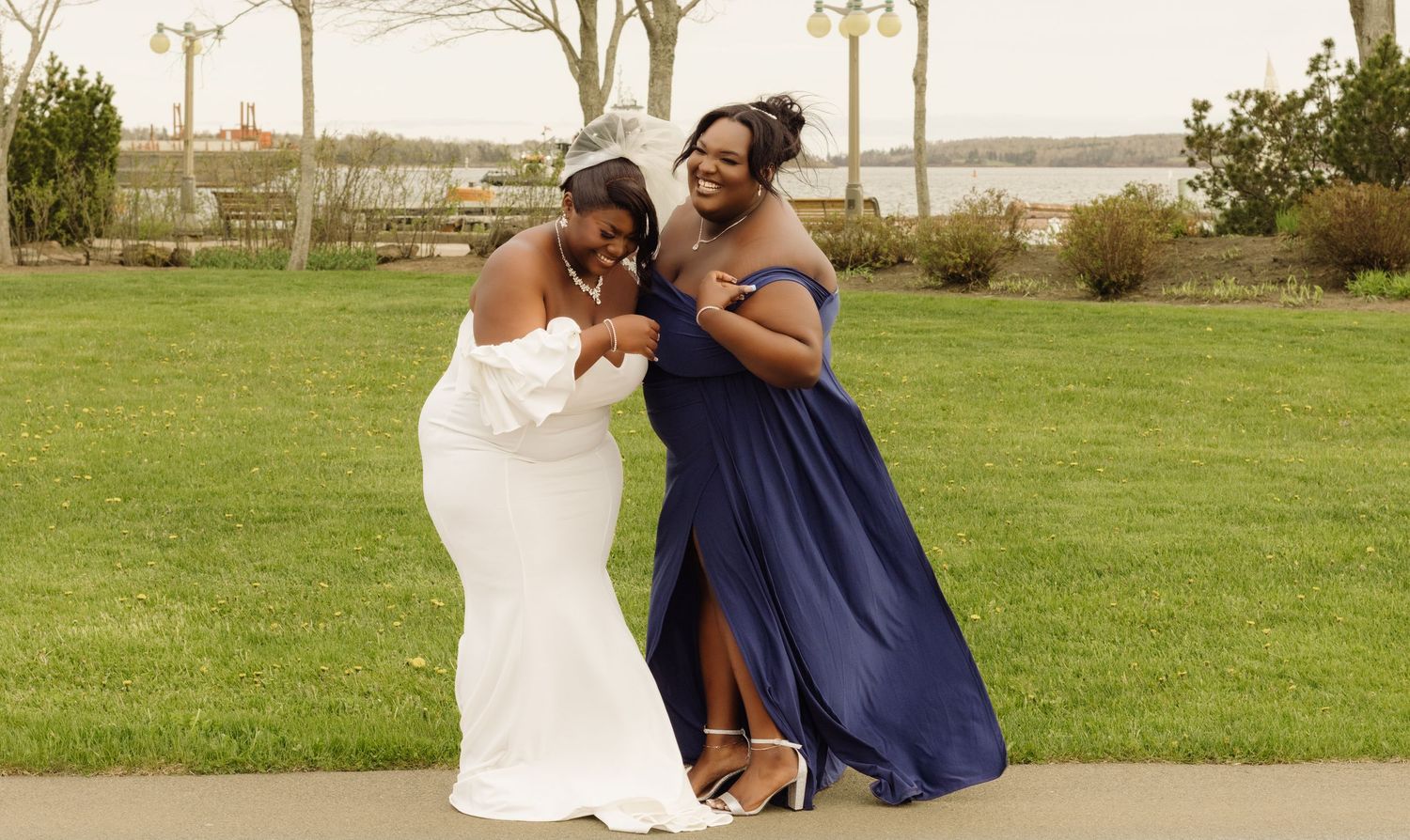 Two people in formal attire sharing a joyful moment on a green lawn by the waterfront.