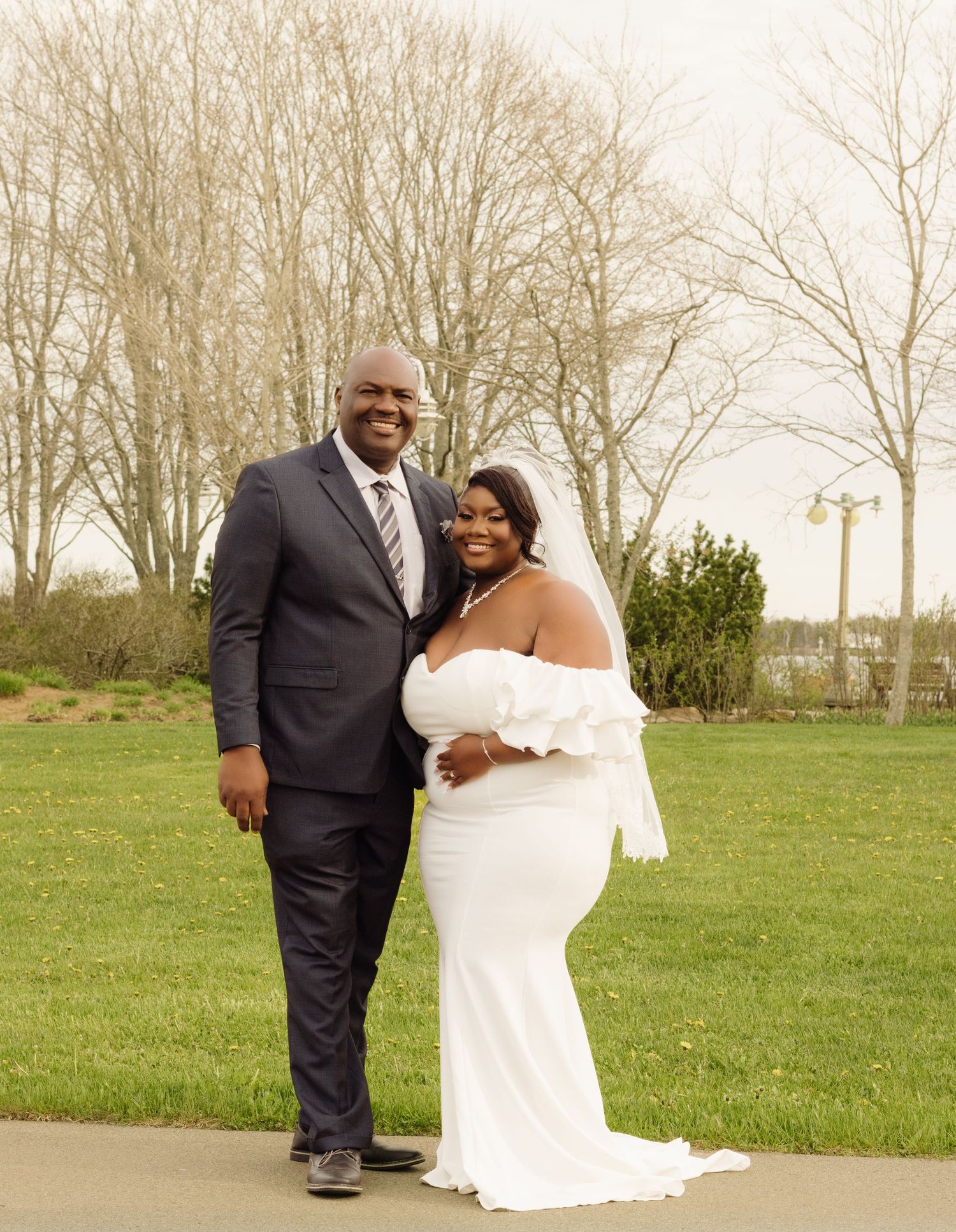 A wedding couple stands together on a green lawn with bare trees in the background on an overcast day.