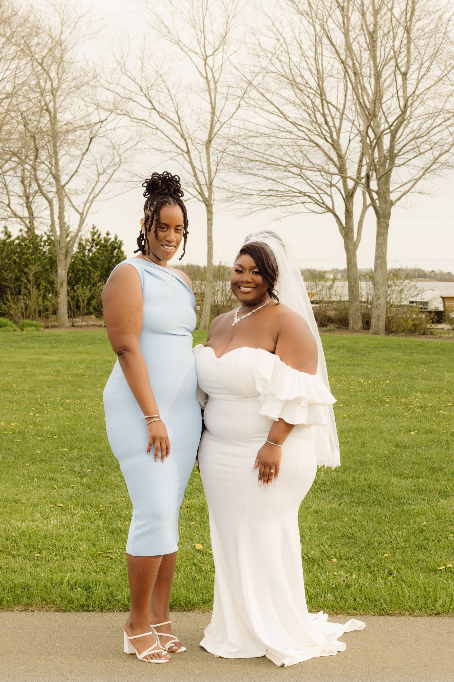 Two women pose together outdoors, one in a light blue dress and one in an elegant white wedding gown.