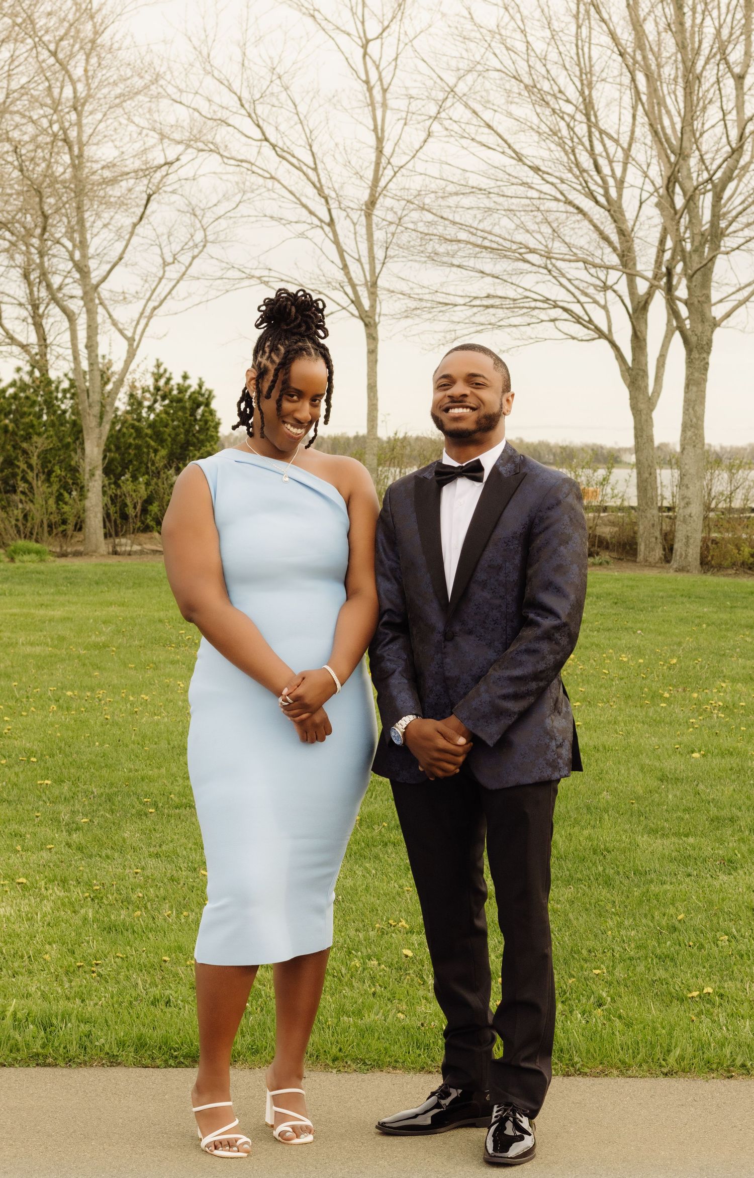 Elegantly dressed couple posing outdoors in formal attire, with her in light blue dress and him in dark suit.