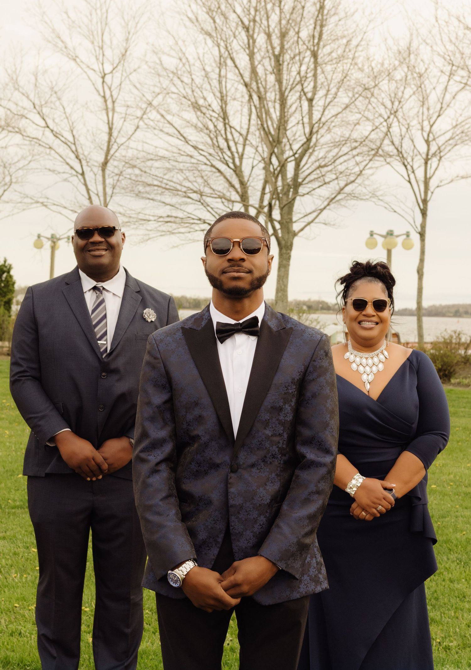 Elegant wedding party in formal attire poses outdoors with bare trees and water in background.