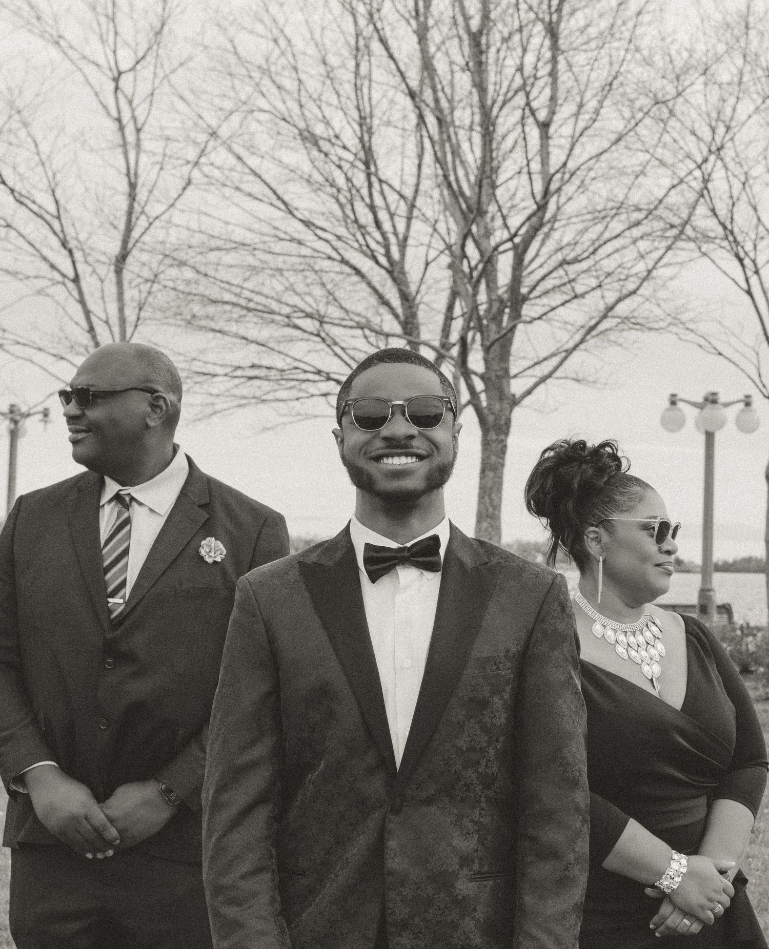 A vintage black and white photograph shows three well-dressed people at a formal event wearing sunglasses and elegant attire.