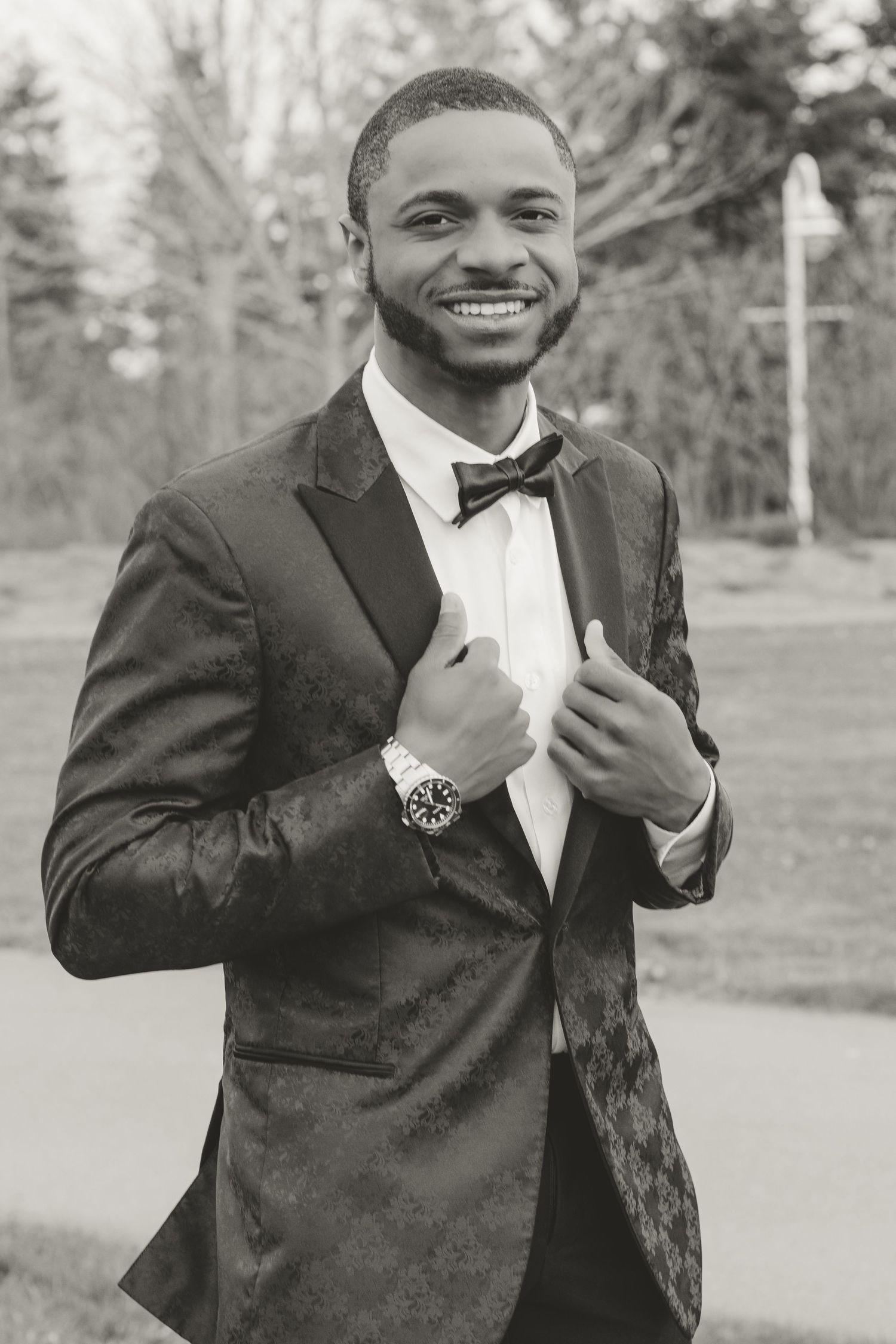 Portrait of a groom in a black tuxedo and bowtie smiling while adjusting his jacket outdoors.