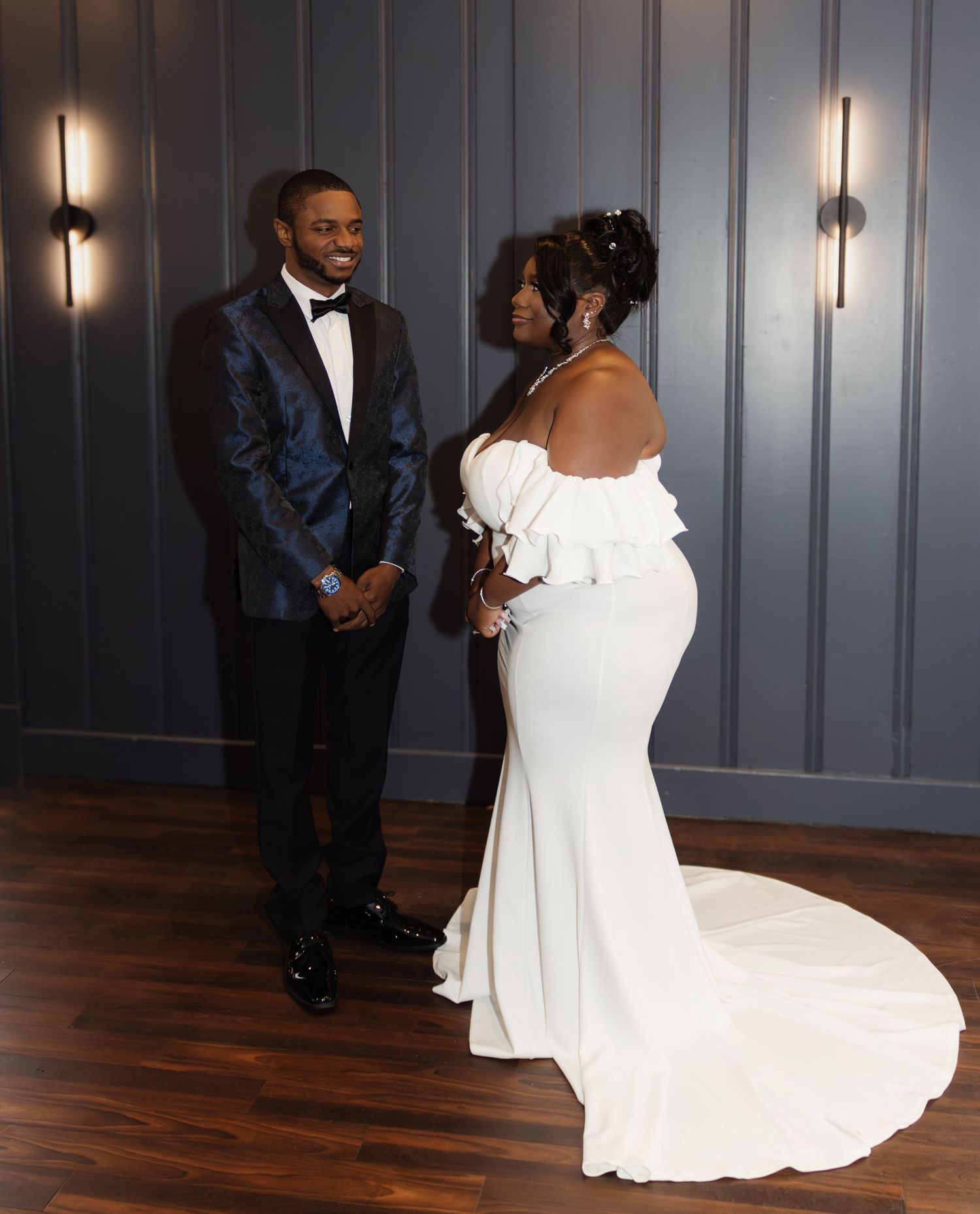 Elegant bride in white off-shoulder gown with train stands with groom wearing navy tuxedo in modern indoor setting.