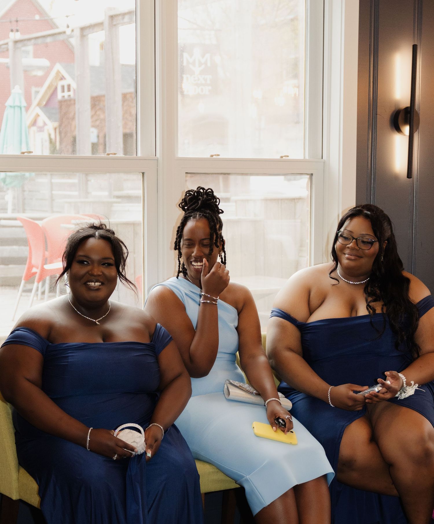 Three bridesmaids in navy blue dresses sit by a sunny window at a wedding celebration.