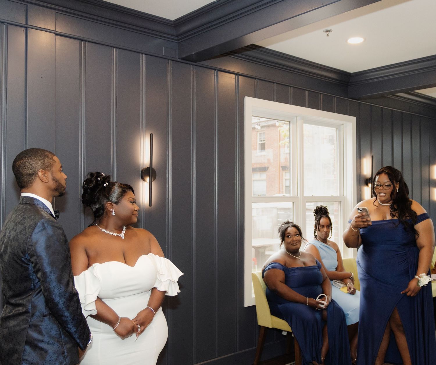 A small wedding party gathers in a room with gray paneled walls and modern sconce lighting fixtures.