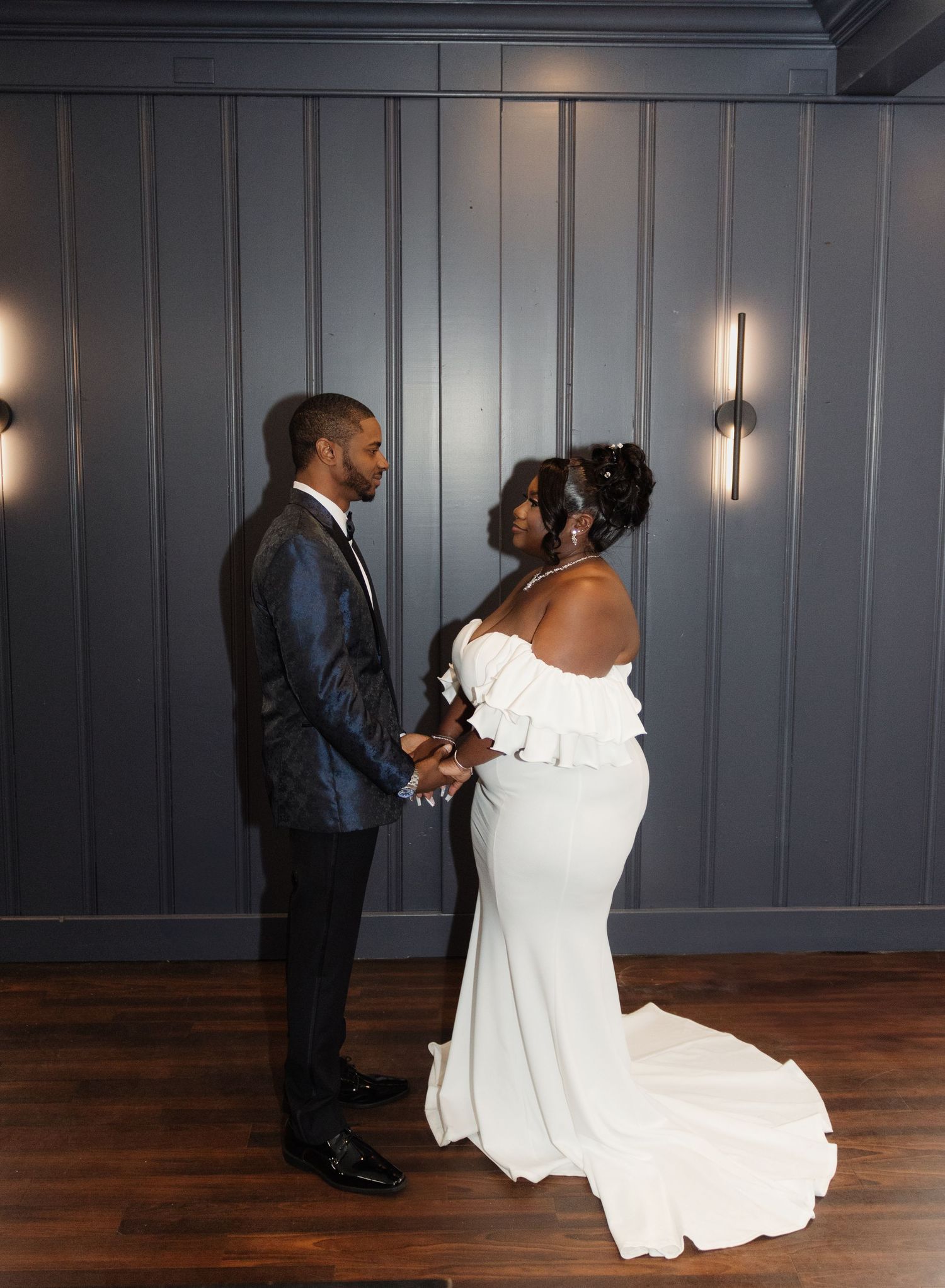 A couple shares an intimate moment in their wedding attire against a grey wall with ambient lighting.