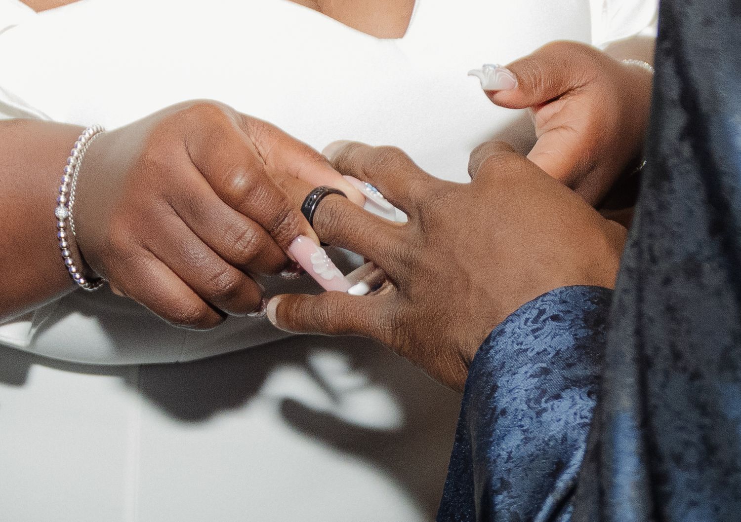 Close up shot of hands exchanging rings during a wedding ceremony with a delicate bracelet and blue fabric visible.