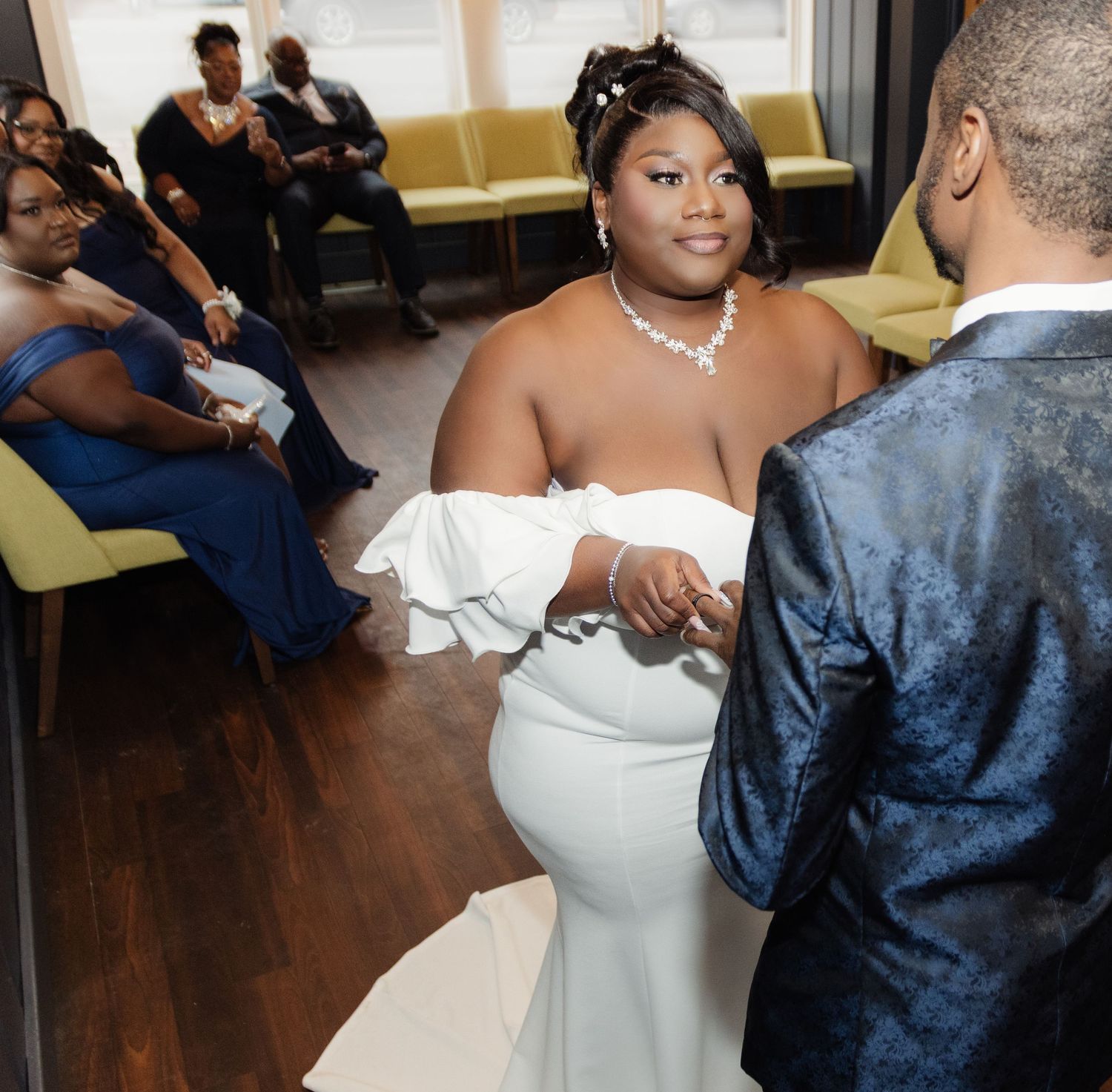 Wedding ceremony in progress with guests seated on yellow chairs in an indoor venue with wooden floors.