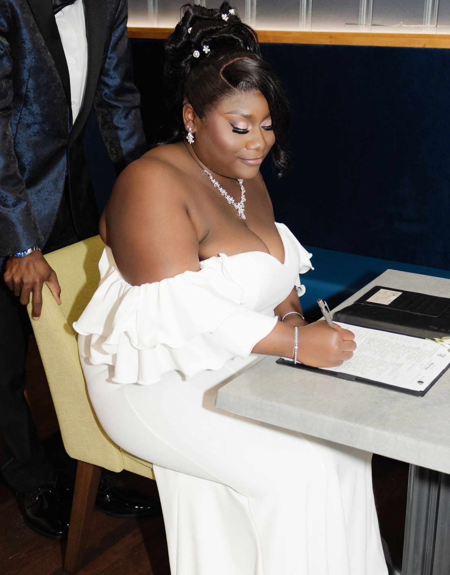 Bride in an elegant white off-shoulder gown signing wedding documents at a table.