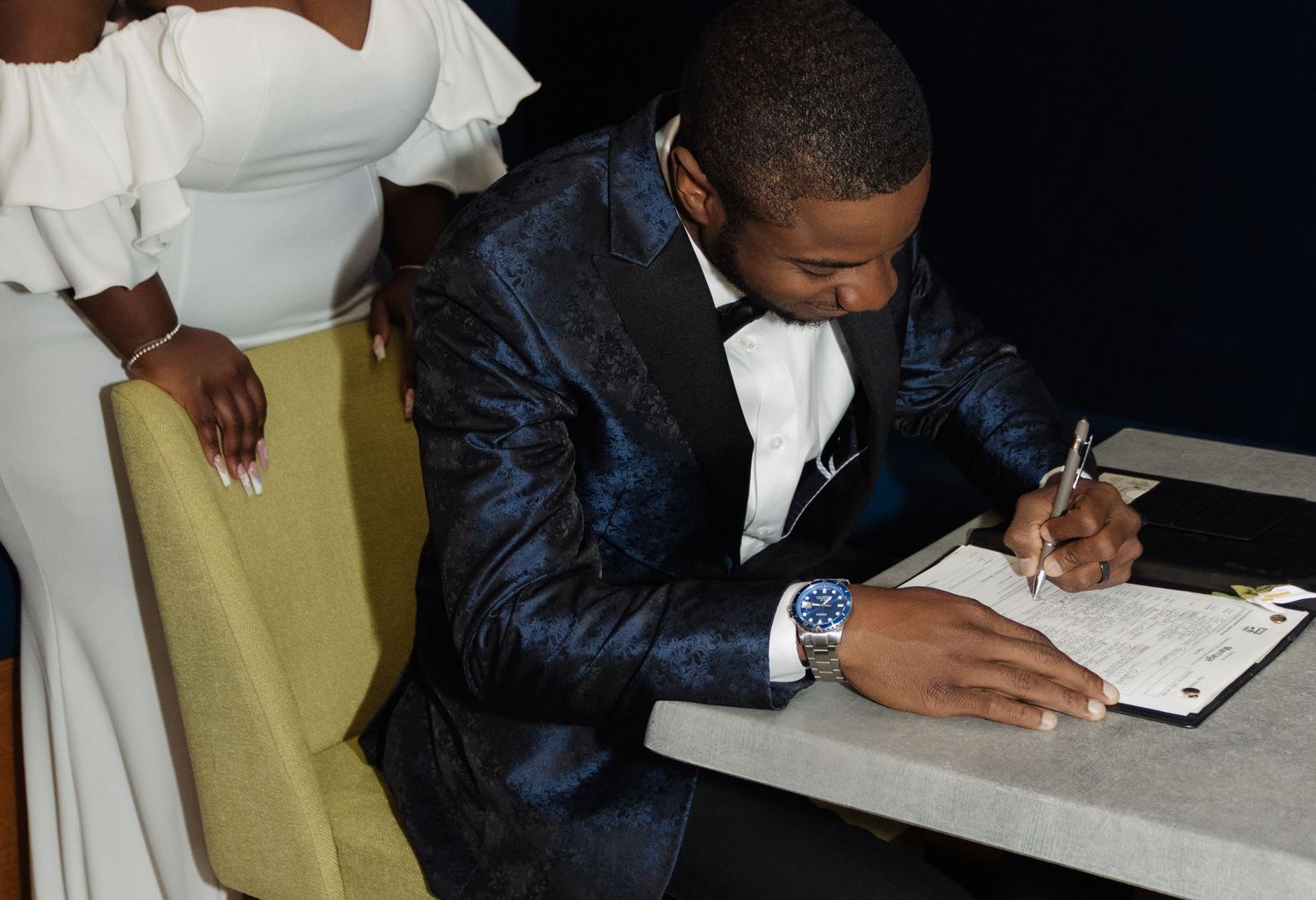 Someone wearing a shiny blue suit and white tie signs a document at a table while another person stands nearby.
