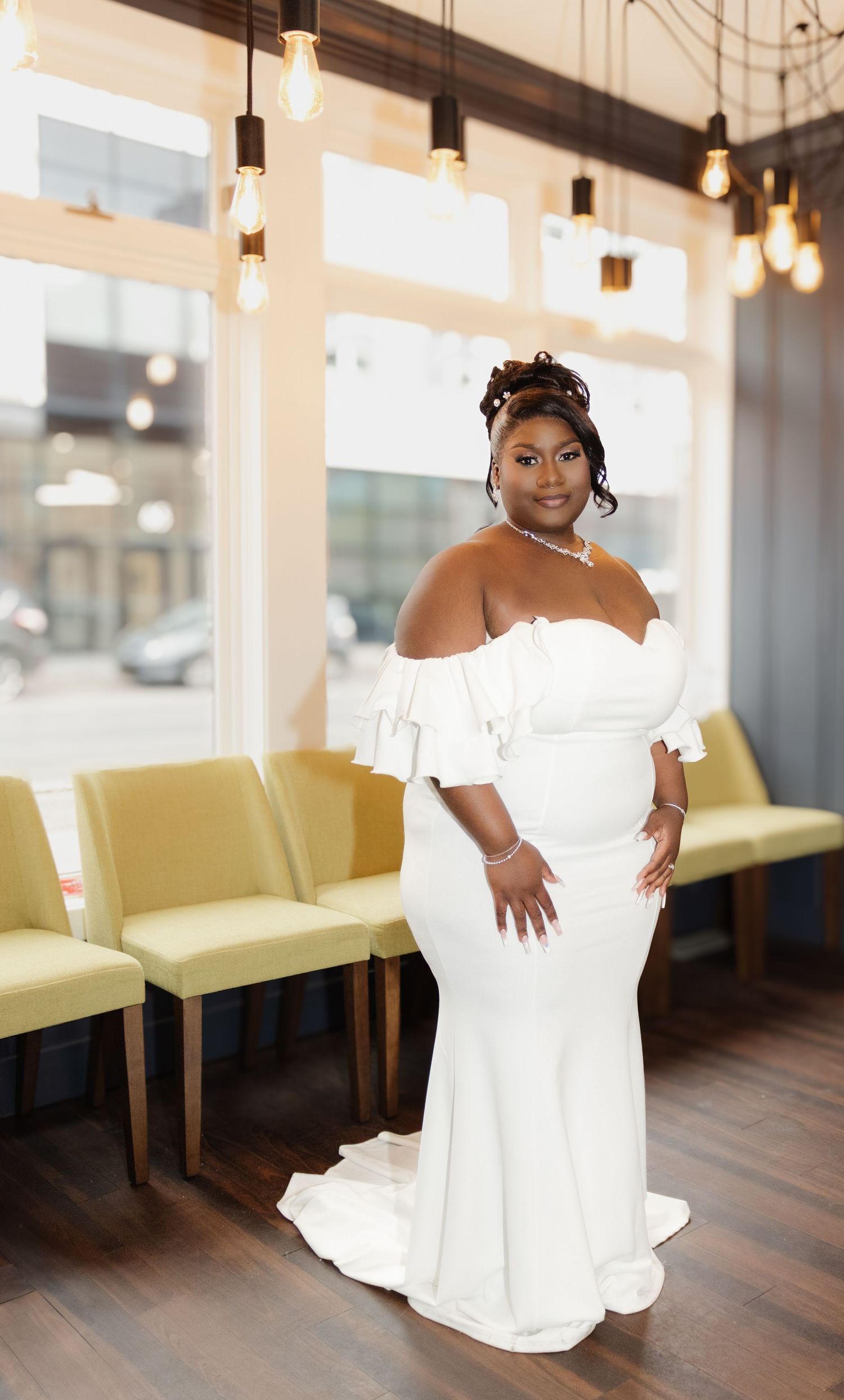 A stunning bride poses in an off-shoulder white wedding gown against a modern interior with pendant lights.