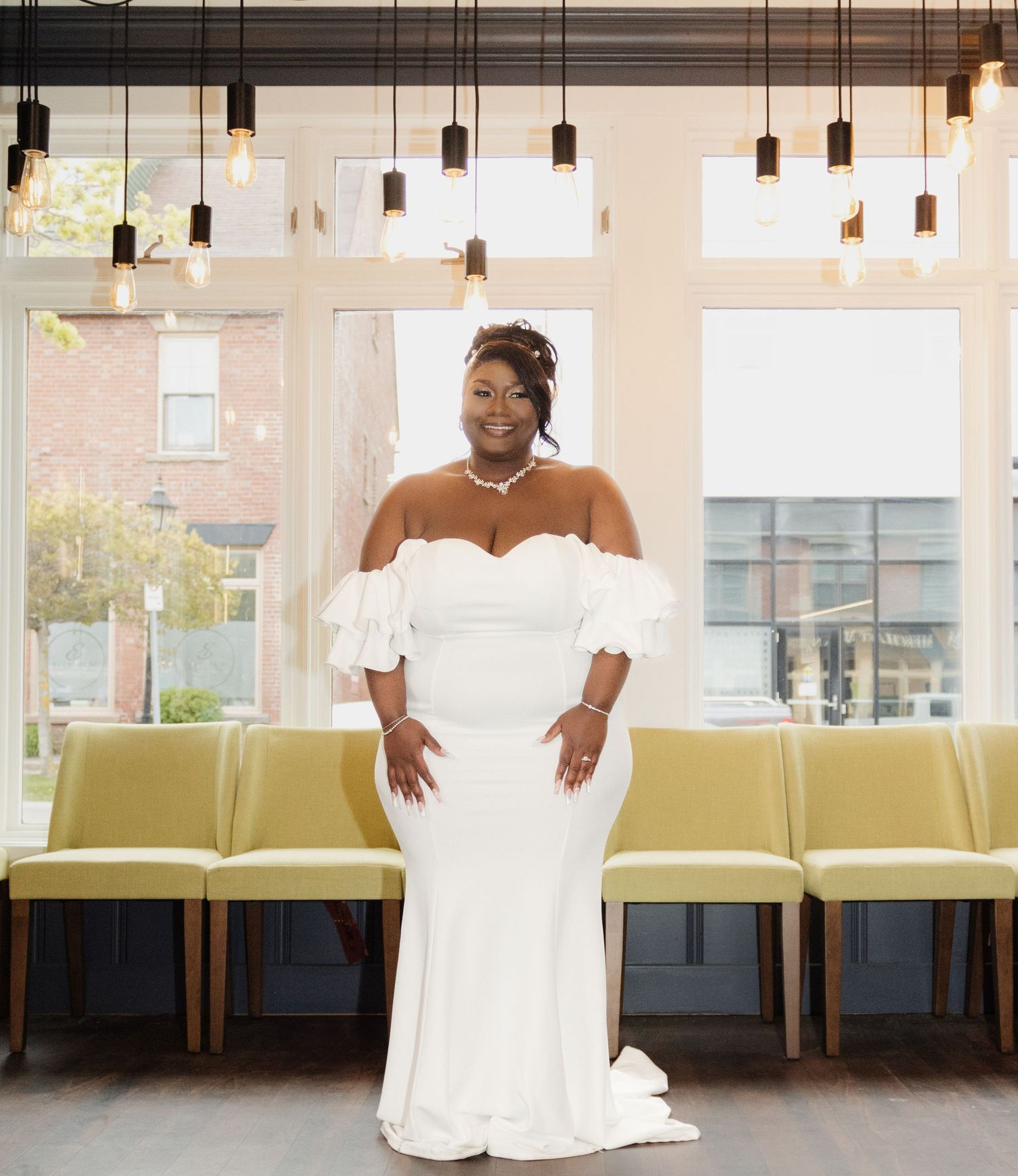 Elegant bride poses in off-shoulder white wedding gown under modern pendant lights in contemporary venue.