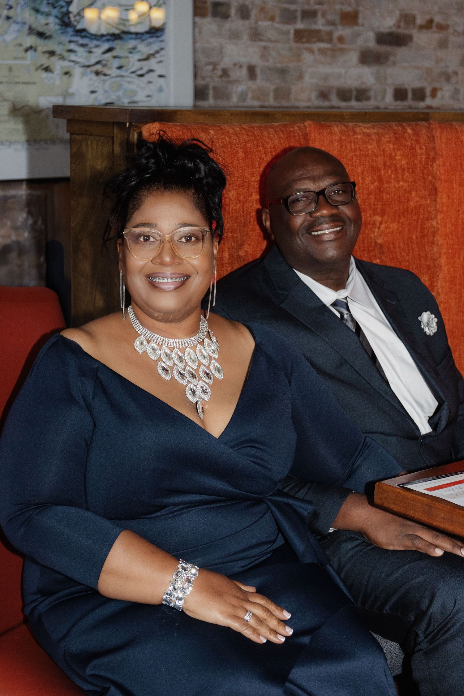 An elegant couple in formal navy attire poses on an orange couch against exposed brick wall.