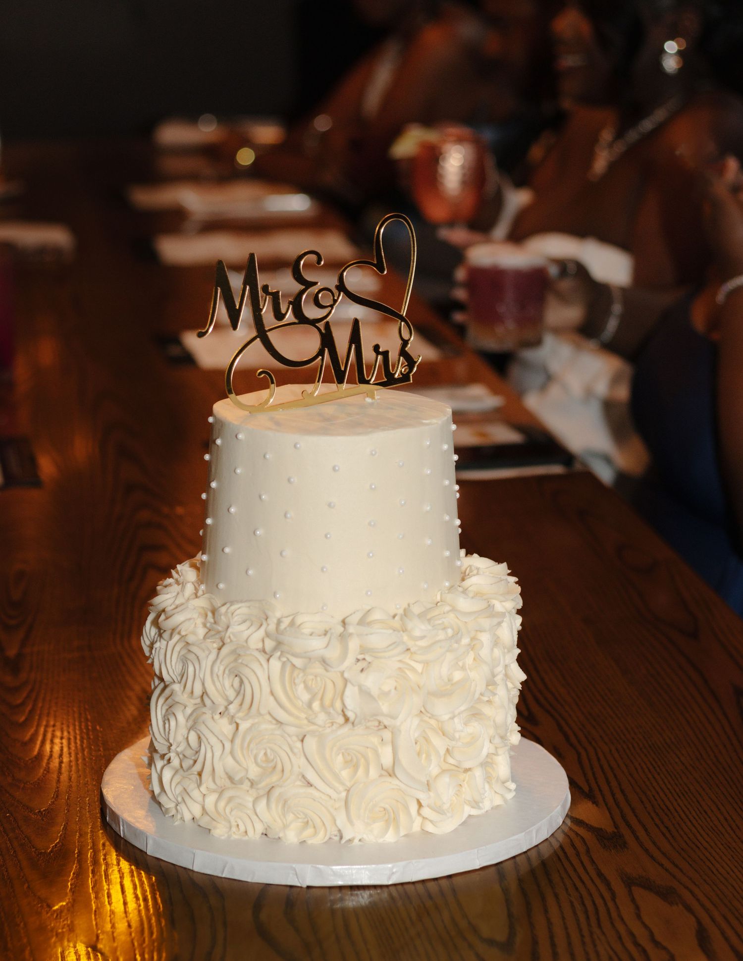 Two-tier white wedding cake with textured frosting and gold 'Mr & Mrs' cake topper on wooden table.