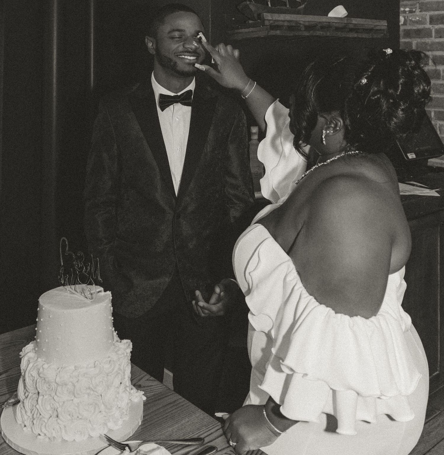 Black and white wedding photo showing a couple laughing together next to their traditional white tiered cake.