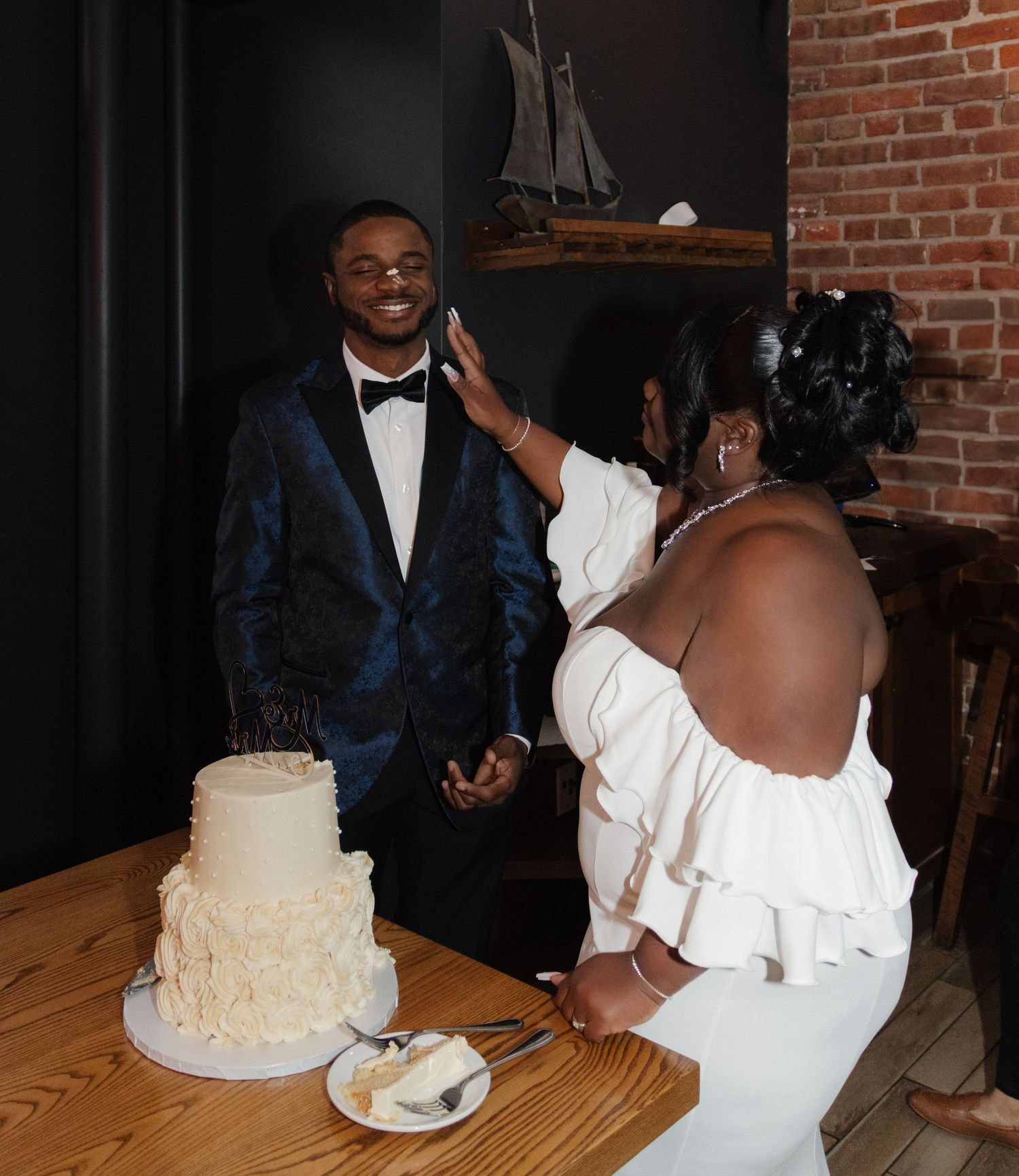 A couple shares a joyful moment beside their tiered white wedding cake in a rustic brick-walled venue.