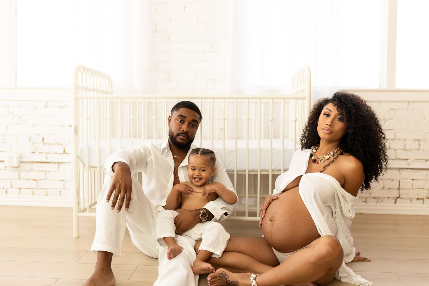 A loving family relaxes on the floor of a bright room with white decor, featuring an expecting parent and a small child.